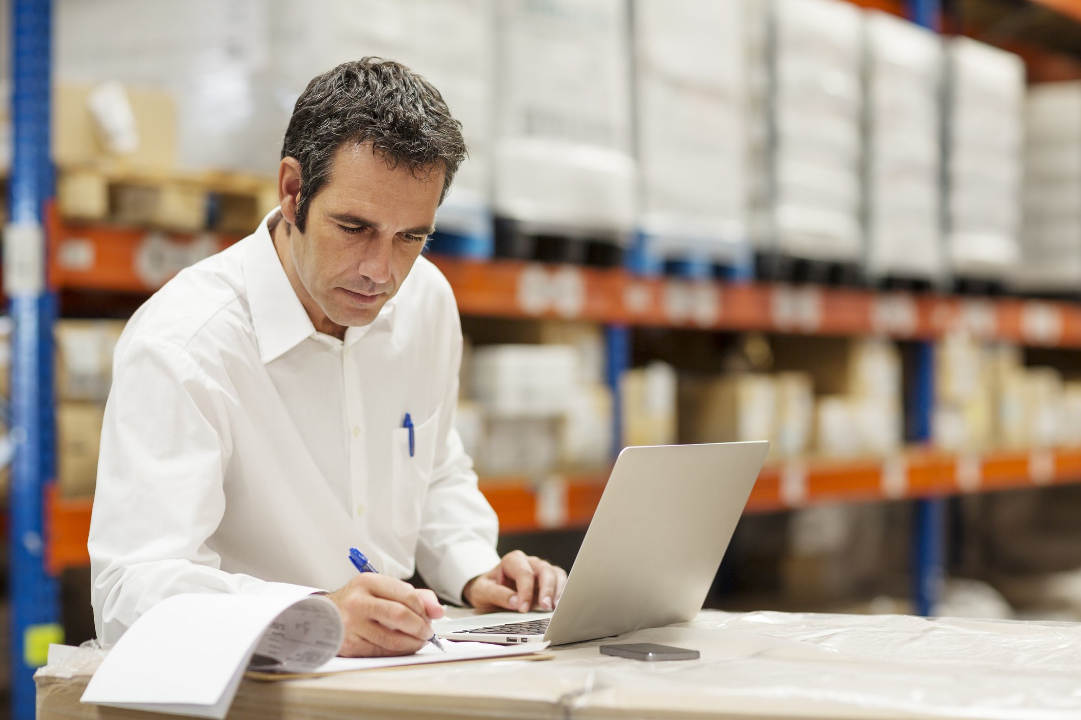 Supervisor doing paperwork while using laptop in warehouse